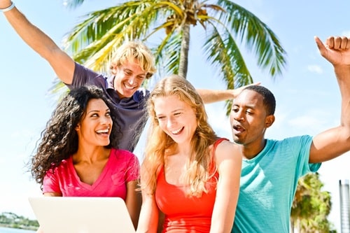 Eine fröhliche Gruppe junger Menschen ist draußen mit einem Laptop, im Hinergrund steht eine Palme